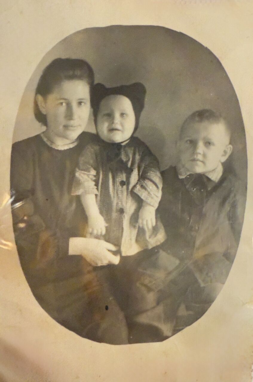 Старая фотография в овальном паспарту: молодая женщина держит на руках малыша в вязаной шапочке с ушками, рядом с ней — мальчик постарше.