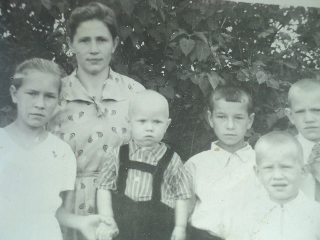 Старая чёрно-белая фотография на фоне листвы: молодая женщина в светлом платье и несколько детей, самый младший — в клетчатой рубашке и тёмном комбинезоне.